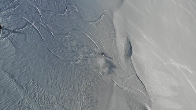 Aerial view of a person skiing downhill in a remote snow-covered mountain with another person standing watching.