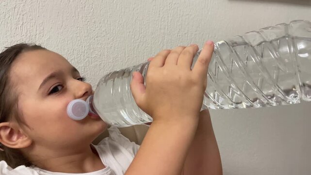 Cute little girl drinking fresh still mineral water from big clear plastic bottle indoors horizontal stock video. High quality 4k footage