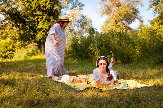 Middle-aged mother wearing straw hat and dress and adult daughter having family rest outdoors in forest area. Sharing, talking, and connecting.