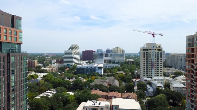 Stepped modern facade overlooks Oak Lawn corridor in Dallas. Red construction crane rises above dense tree shaded multi family enclaves beside beige corporate office towers, Texas