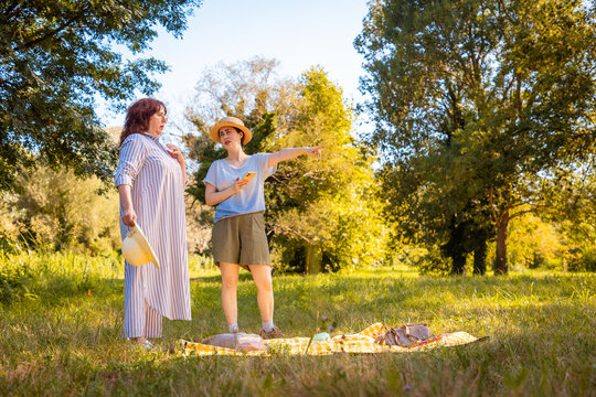 Adult daughter and mother enjoy a summer picnic in a park surrounded by grass and trees engaging in playful leisure and family quality time.