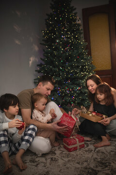 A large family of three children, mom and dad, are celebrating New Year's and Christmas under the Christmas tree at home, opening presents. A quiet, cozy family evening.