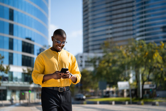 Casual style, Black smiling businessman walking, using smartphone phone, texting, near glass office building. Mid age African american business man works as Investor, reading news app, modern living.