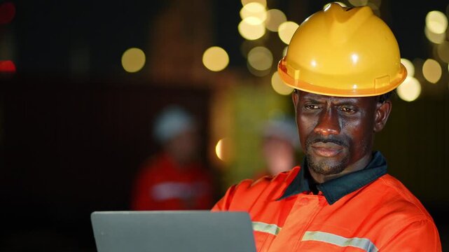 Black male technician wearing safety uniform typing on portable laptop computer during outdoor night inspection task at industrial oil refinery plant