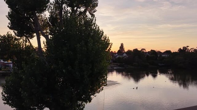 Sideways and forward drone tracking shot emerging from behind a tree to reveal the sunset over the famous lake in Raphael Park, Romford