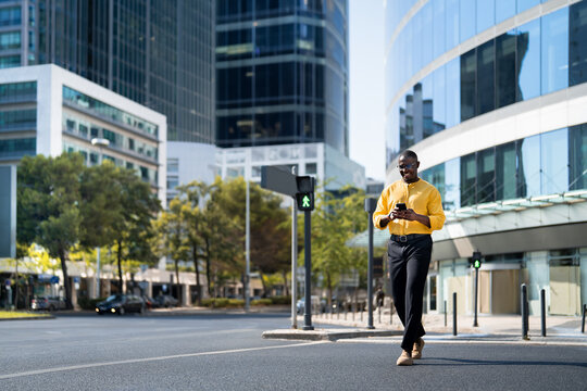 Fullbody, African american smiling businessman walking, using smartphone phone, texting, near glass office building. Mid age Black business man works as Account Executive, active. Copy space.