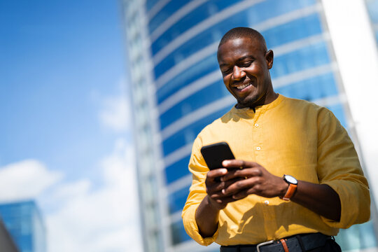 Standing, African american smiling businessman using smartphone phone, texting, near glass office building. Modern Black business man works as Manager, chatting with AI assistant, staying connected.