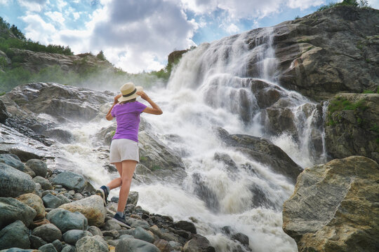 Back view of a woman holding her straw hat while looking at a powerful rushing mountain waterfall. People and nature landscape