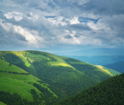 Sunlight breaks through dark storm clouds onto vast rolling green hills with a small river valley. Dramatic alpine landscape