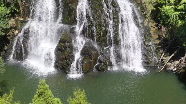 Water cascades over rocky cliffs into a green pool surrounded by lush forest, North Island, New Zealand