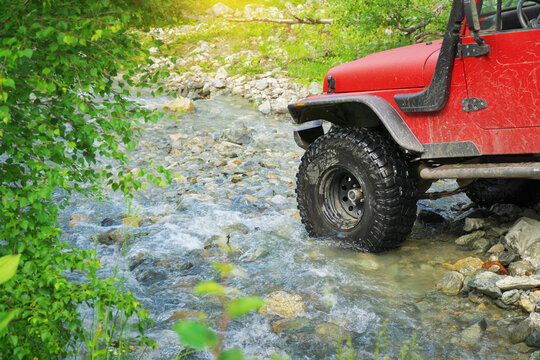 Close up of a large mud tire on a red offroad vehicle crossing a shallow mountain river by green trees. Automotive detail image suits