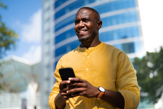 African american smiling businessman using smartphone phone, texting, near glass office building, standing. Mature Black business man works as Trader, chatting with friends, tech savvy lifestyle.