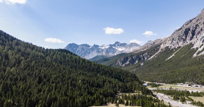 Passo del Forno forested valley pulls back to reveal distant alpine peaks under clear midday skies.