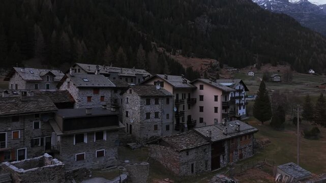Old stone village houses sit below dark forested slopes at dusk in Alpe Ventina, Valmalenco.