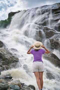 Back view of a woman holding her straw hat while looking at a powerful rushing mountain waterfall. People and nature landscape design