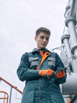 Low angle view of young male technician adjusting safety gloves. Industrial worker in teal and orange uniform at refinery. Maintenance and safety concept