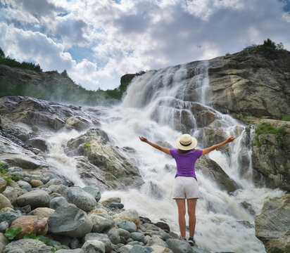 Woman in straw hat raises hands by a massive powerful waterfall cascading down rocky mountain cliffs. Inspiring freedom concept