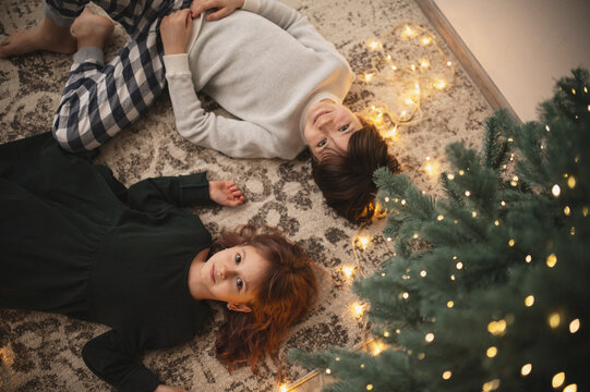 New Year's card with garlands, top view. Brother and sister are lying under the Christmas tree on the floor on a fluffy carpet. Bokeh. Christmas atmosphere.