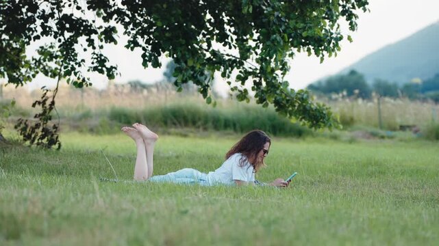 Young woman lying on her stomach on the grass and using a smartphone under a large tree, enjoying a relaxed outdoor moment in nature.