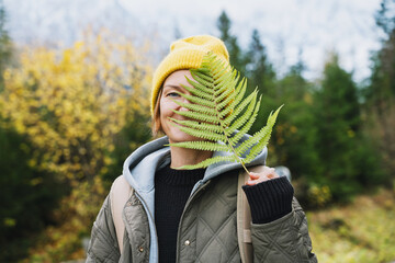 A smiling young woman in a yellow beanie playfully holds a green fern leaf against an autumn forest background. Joyful outdoor lifestyle concept radiates happiness and a deep connection with nature. © nataliaderiabina