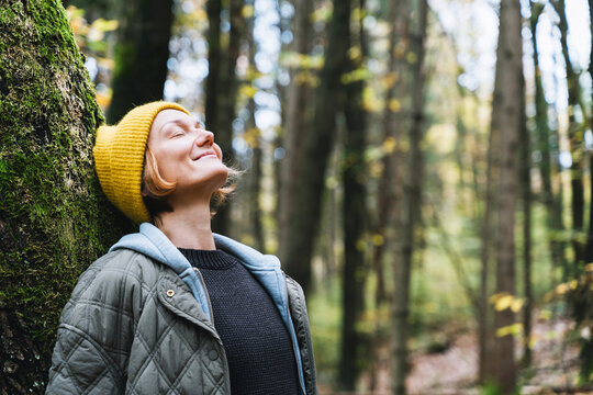 A young woman in a yellow beanie leans comfortably against a massive tree trunk in a quiet autumn forest. Relaxing nature connection concept emphasizes the grounding 