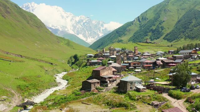Ushguli settlement approaches in forward aerial view beside the Enguri River and alpine slopes, supporting cultural tourism, rural geography, medieval heritage and mountain travel promotion in Georgia