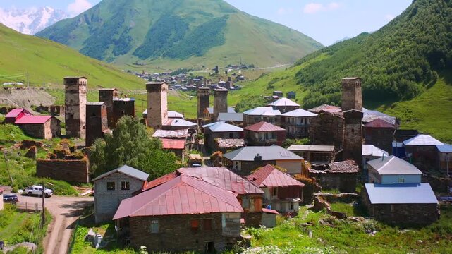 Drone advances above Chazhashi homes in aerial view toward clustered Svan towers and mountain fields, supporting medieval architecture, heritage preservation and Upper Svaneti village tourism.