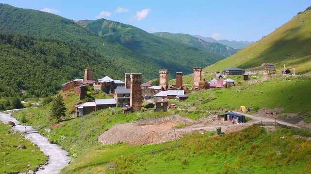 Drone advances toward Chazhashi towers, aerial view across stone homes and green slopes in Ushguli, supporting medieval heritage, rural tourism, architectural research, Georgian cultural travel media