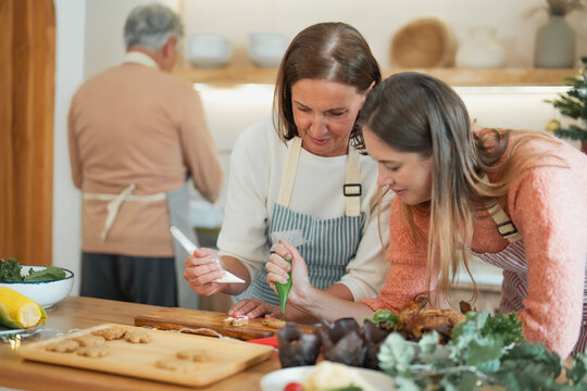 Mother, adult daughter decorating homemade Christmas cookies with colorful icing while senior father prepares food in background during family holiday gathering.