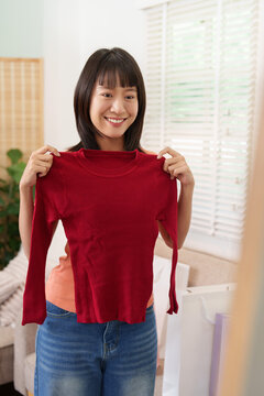 Happy Asian woman holding red sweater for online shopping clothing try on at home
