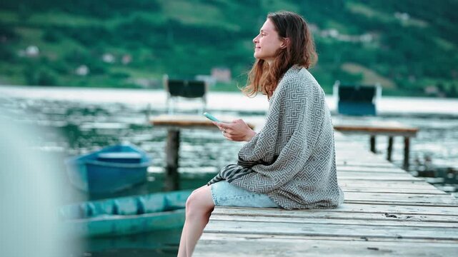 Young woman wrapped in a warm blanket sitting on a wooden pier by a scenic mountain lake and using a smartphone, enjoying peaceful nature while staying connected during her travels.