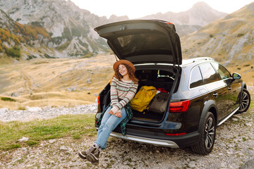 Woman in hat sits on edge of car's trunk, admiring mountain scenery. Woman on road trip stands next to car with hiking backpacks in the trunk. Concepts of relaxation, travel, and freedom. © maxbelchenko