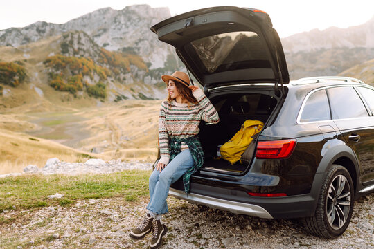 Woman in hat sits on edge of car's trunk, admiring mountain scenery. Woman on road trip stands next to car with hiking backpacks in the trunk. Concepts of relaxation, travel, and freedom.