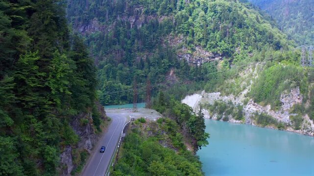 Drone flies backward above Enguri Reservoir road along turquoise water and wooded cliffs in Svaneti, Georgia, supporting scenic travel, transport mapping, infrastructure media and regional tourism.