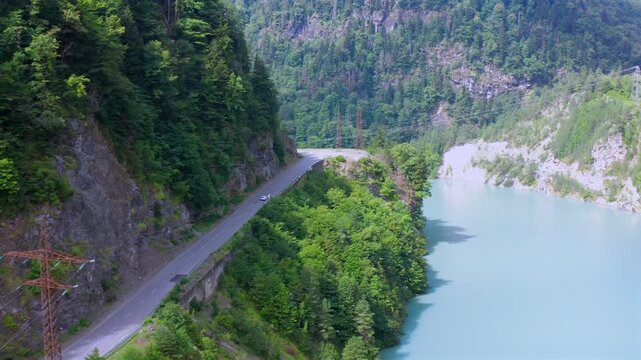 Drone flies forward above Enguri Reservoir road as it curves between forested cliffs and turquoise water, supporting Georgian road tourism, regional mobility, landscape geography and travel promotion.