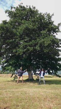Funny video of two female travelers wearing bucket hats dancing playfully in front of a large tree, capturing friendship, humor, carefree travel and fun outdoor moments.