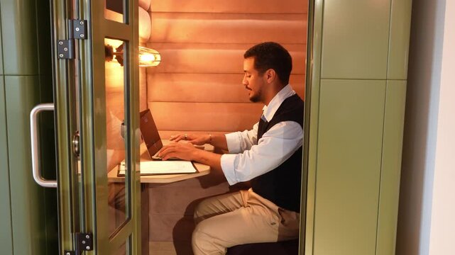 Confident male professional typing on a laptop computer inside a private soundproof booth in a contemporary coworking space, then smiling