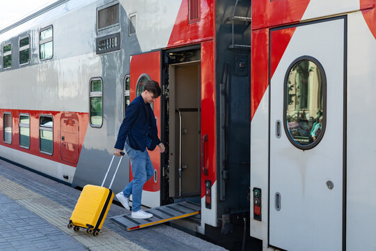 Man carrying a yellow suitcase walks up the ramp to board a double-decker train at a modern railway station, with the train's exterior featuring red and gray colors