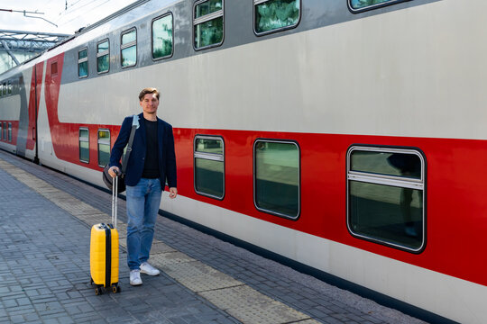 Young man with yellow suitcase walks beside a red and gray train at a railway station, preparing for travel on a sunny day. Business trip and vacation concept.