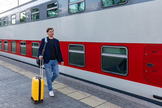 Young man with yellow suitcase walks beside a red and gray train at a railway station, preparing for travel on a sunny day. Business trip and vacation concept.
