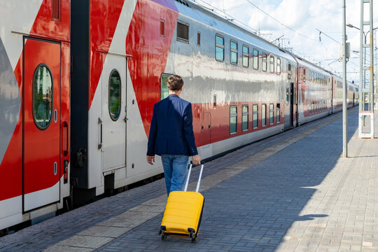 Rear view young man in a blue blazer and jeans pulls a yellow suitcase while walking next to a modern train at a railway station, highlighting urban travel and the dynamic atmosphere of transportation