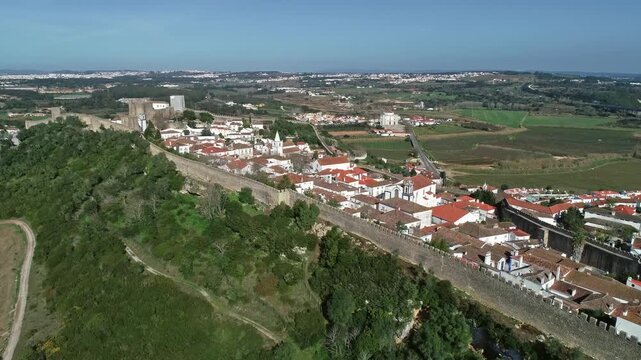 Aerial view of the historic walled town of Obidos with its medieval fortifications, red-tiled rooftops, and white buildings surrounded by green countryside in Obidos, Leiria District, Portugal.
