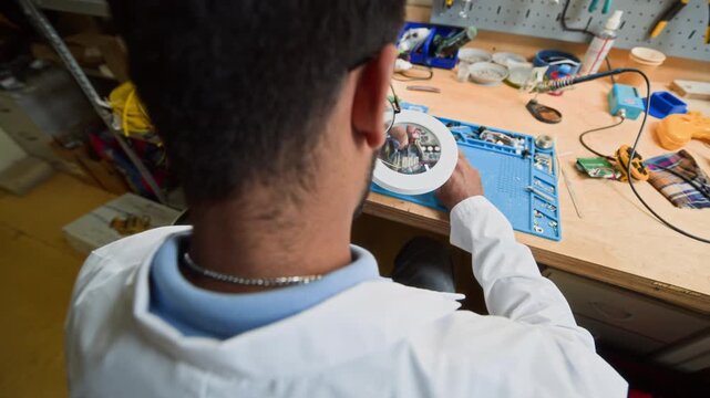 Indian young adult male engineer examining robotic chassis under magnifying glass lamp in laboratory. High angle arc shot ideal for promoting STEM education and tech innovation
