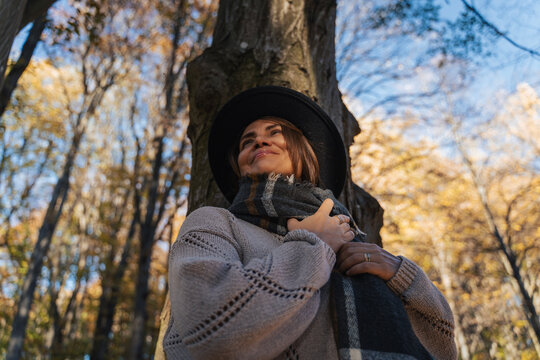 Low angle view of a smiling mid-adult woman in a felt hat by a tree in autumn forest.