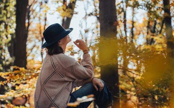 Mid-adult woman sitting on a log in autumn forest with a felt hat pulled down over her eyes.
