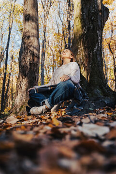 Vertical portrait of a peaceful mid-adult woman sitting in a lotus position by a tree trunk in autumn forest.