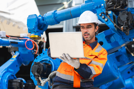 Robotics engineer using laptop to program industrial robot arm, automation engineer working with computer at robotic manufacturing plant, factory technician operating robotic machine with laptop com