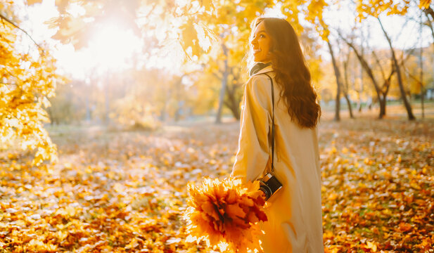 Happy woman in light-colored cape is having fun in autumn park at sunset. Woman with a camera and a bouquet of yellow leaves is enjoying a walk outdoors. Autumn mood. Lifestyle.