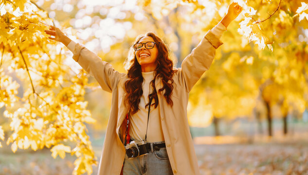 Happy woman in light-colored cape is having fun in autumn park at sunset. Woman with a camera and a bouquet of yellow leaves is enjoying a walk outdoors. Autumn mood. Lifestyle.
