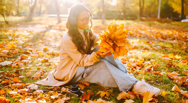 Happy woman in light-colored cape is having fun in autumn park at sunset. Woman with a camera and a bouquet of yellow leaves is enjoying a walk outdoors. Autumn mood. Lifestyle.